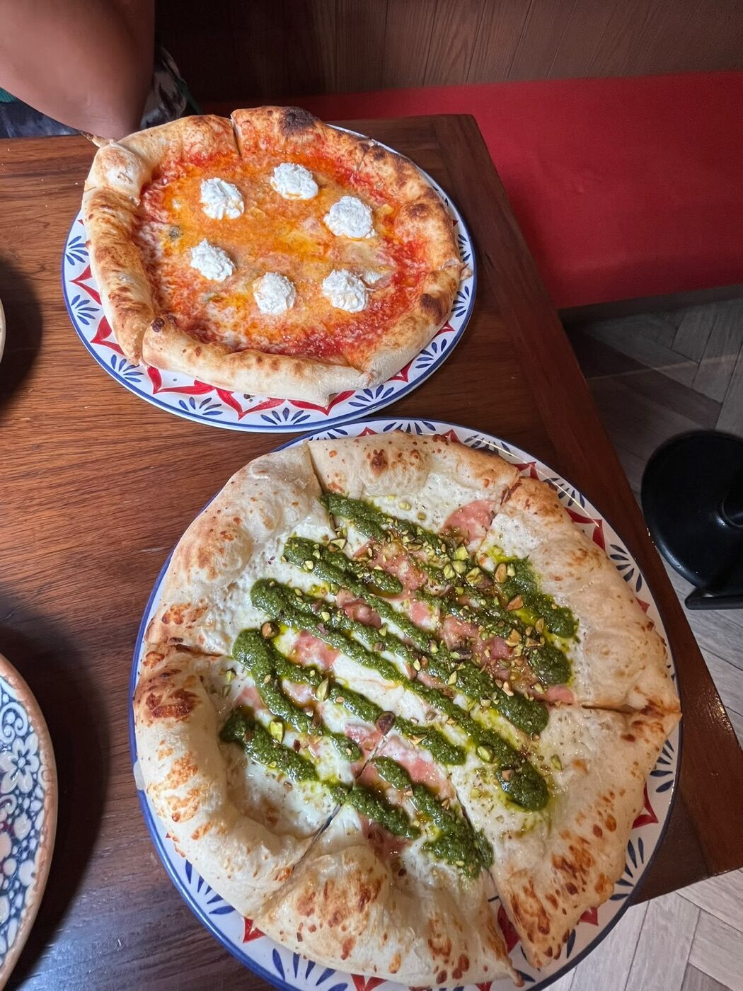 Dos pizzas personales en Marcello: una roja con bolitas de ricotta y otra blanca con mortadela, pesto y pistache, en platos de cerámica azul y roja
