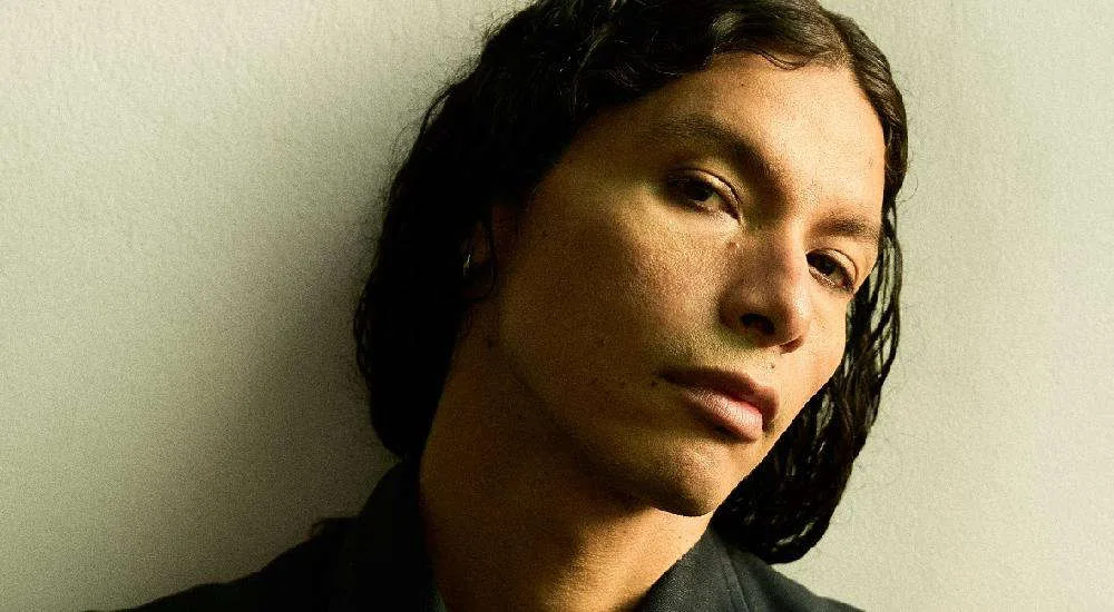 Retrato de A.Chal, cantante y productor peruano, de cabello largo y negro, frente a una pared clara