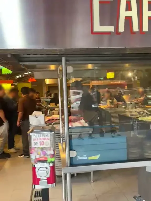 Interior de Asaderos Orozco visto desde la entrada, con la barra de la parrilla, gente formada y una máquina de chicles