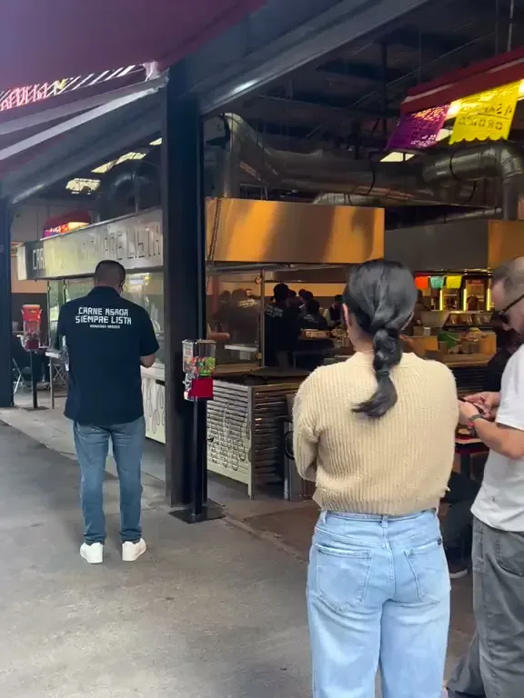 Fila de personas en la entrada de Asaderos Orozco, con el salón tipo bodega y papel picado al fondo