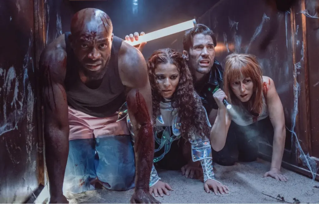 Escena de Bitten Soon: cuatro personajes heridos avanzan a gatas por un túnel estrecho y oscuro, uno de ellos con una lámpara