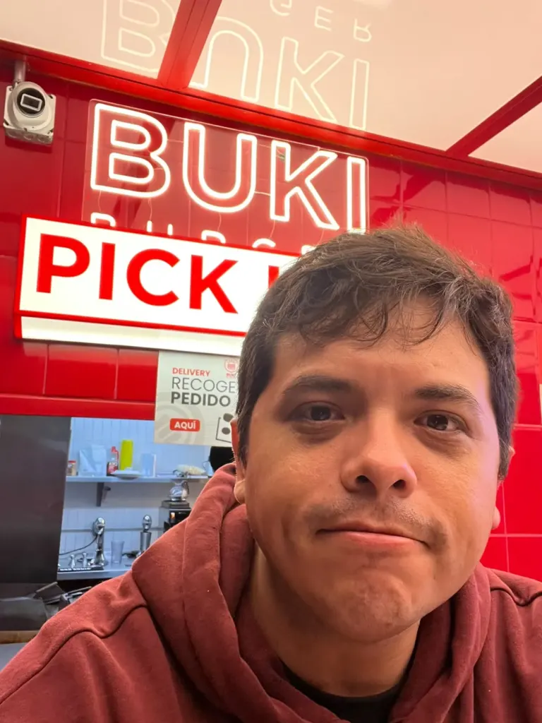 José G., autor de A dónde viajar, frente al mostrador rojo de Buki Burger con el letrero de neón y el cartel de Pick up