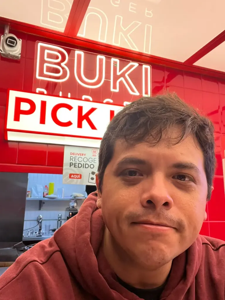 José G. en Buki Burger Roma Norte con sudadera guinda frente al neón rojo de la marca y el mostrador de pedidos