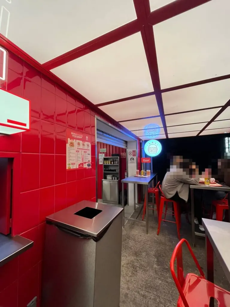 Interior de Buki Burger con azulejo rojo, techo de luz blanca con vigas rojas, bancos rojos y una mesa alta de acero junto a la entrada