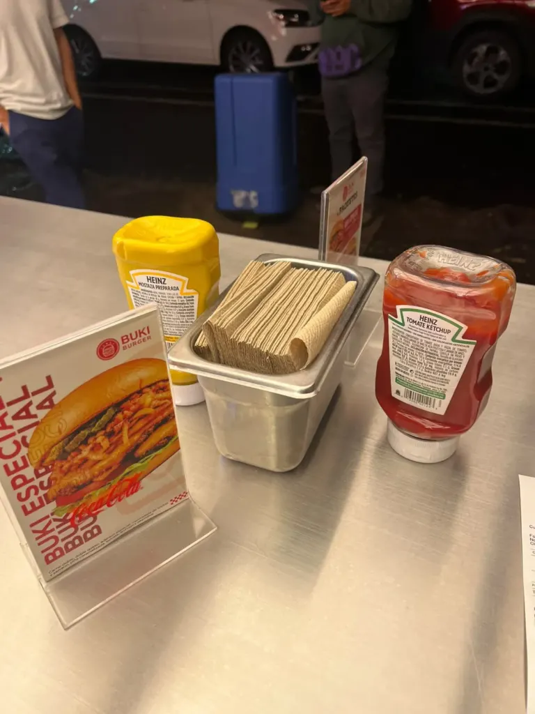 Mesa de acero de Buki Burger con mostaza y cátsup Heinz, servilletero metálico y un cartel de la hamburguesa Buki Especial