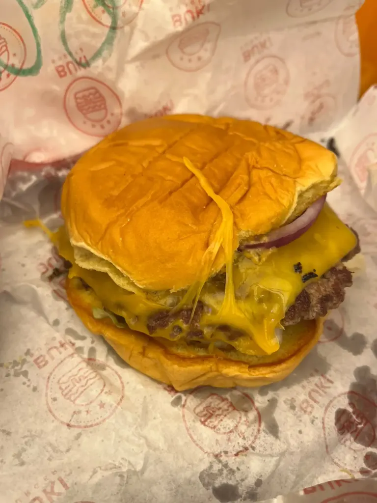 Smash burger de Buki Burger con queso cheddar derretido escurriendo, cebolla morada y pan brioche sobre papel con el logo de la marca