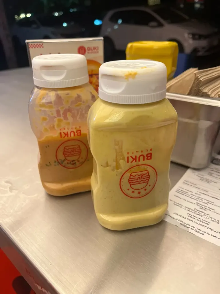 Dos botellas de salsa de la casa de Buki Burger, una anaranjada y otra amarilla cremosa, sobre la mesa de acero