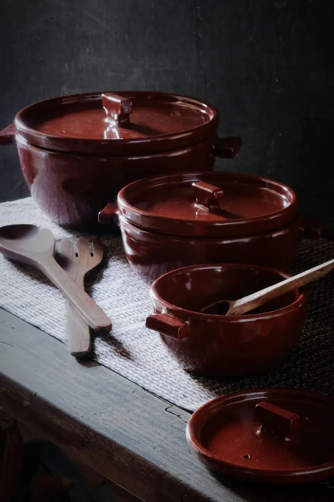Mesa de madera oscura con cazuelas de barro vidriado en rojo, una jarra de vidrio y utensilios, montadas como un bodegón sobre fondo negro, en el estilo de Casa Onora