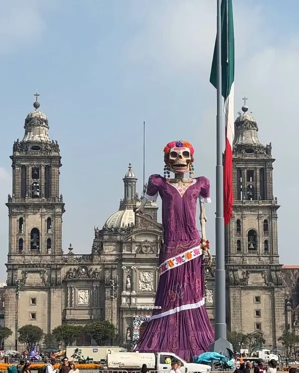 Catrina gigante de 32 metros con vestido morado y flores en la cabeza frente a la Catedral Metropolitana en el Zócalo de la Ciudad de México, junto a la asta bandera