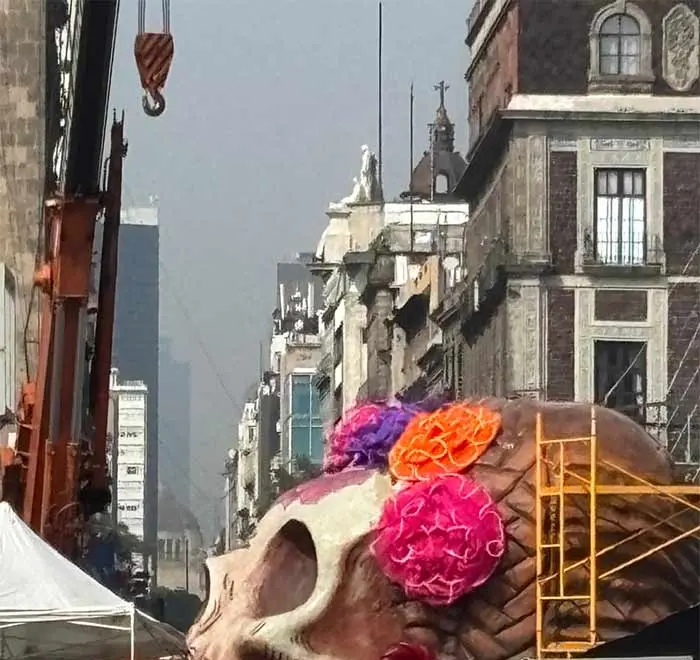 Cabeza gigante de la Catrina del Zócalo con flores rosas, moradas y naranjas, en el suelo antes de ser izada, con una grúa y los edificios del Centro Histórico al fondo