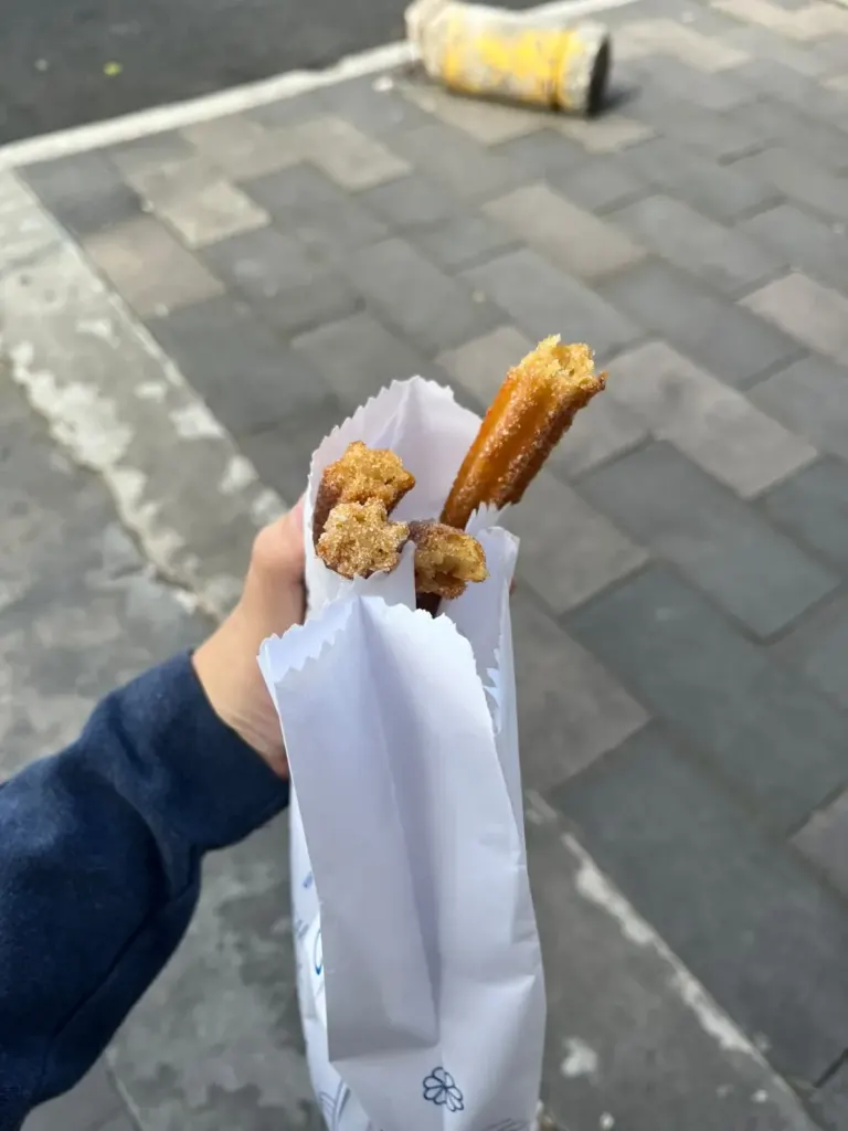 Mano sosteniendo una bolsa de papel de Churrería El Moro con dos churros con azúcar en la banqueta