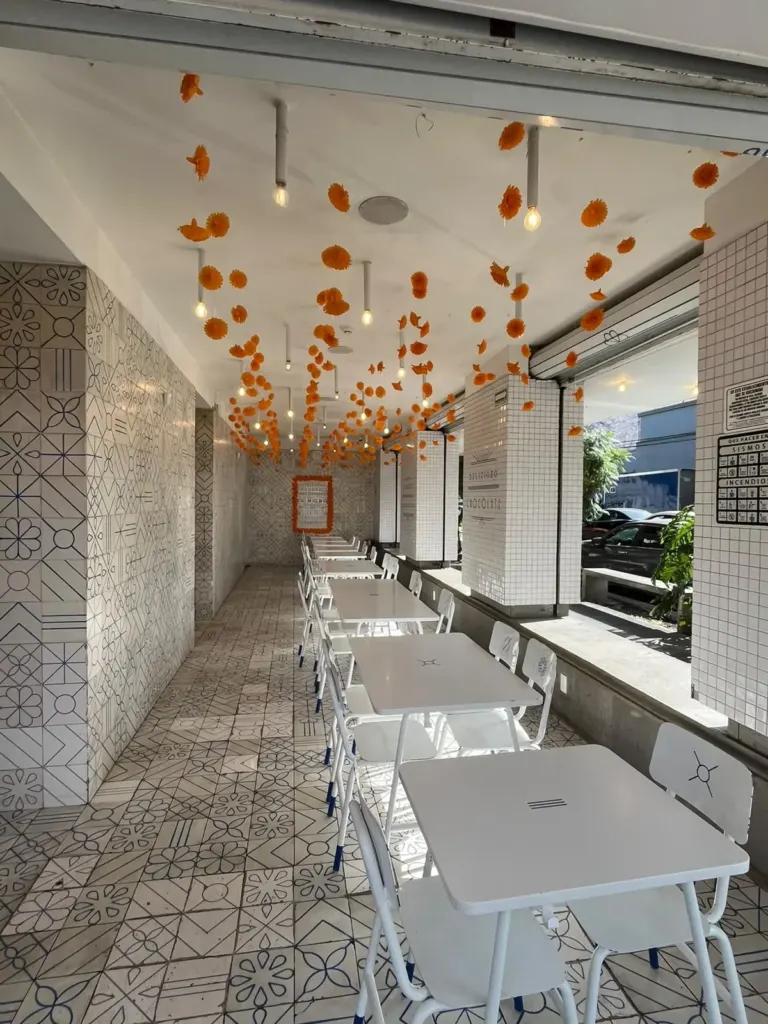 Interior de El Moro Roma con mesas blancas, azulejo y flores de cempasúchil colgando del techo