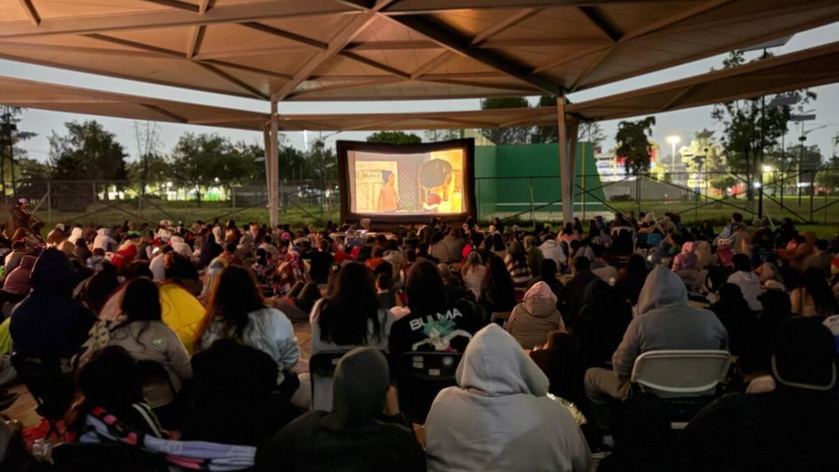 Público sentado bajo una techumbre viendo una película en pantalla al aire libre en el Bosque de San Juan de Aragón al anochecer