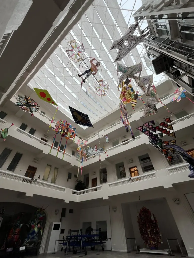 Patio central del Museo de Arte Popular visto desde abajo, con tragaluz de cristal, balcones blancos en varios pisos y papalotes de colores colgando
