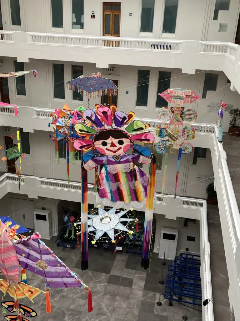 Muñeca tipo Lele gigante con trenzas y vestido de rayas de colores colgada en el patio del Museo de Arte Popular, con una estrella blanca y papalotes alrededor