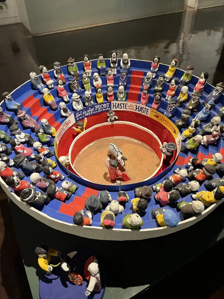 Plaza de toros en miniatura con decenas de calaveras de colores en las gradas, un torero y un toro esqueleto en el ruedo y anuncios pintados de Sal de uvas Picot y Haste