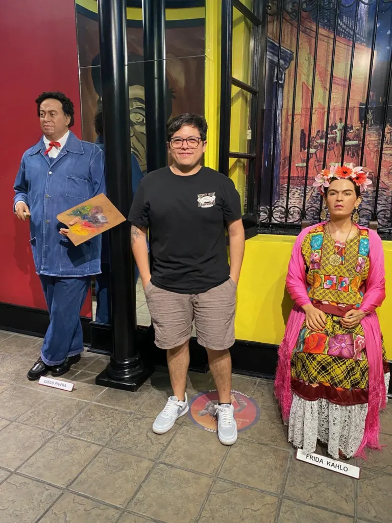 Figuras de cera de Diego Rivera con overol de mezclilla y paleta de pintor y de Frida Kahlo con vestido de tehuana y flores en el cabello, con un visitante en medio