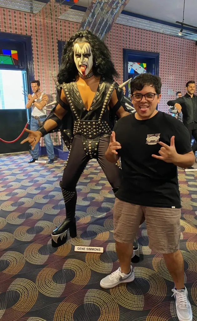 Figura de cera de Gene Simmons de Kiss con maquillaje blanco y negro y traje de cuero con estoperoles, junto a un visitante haciendo cuernos con las manos, en una sala alfombrada