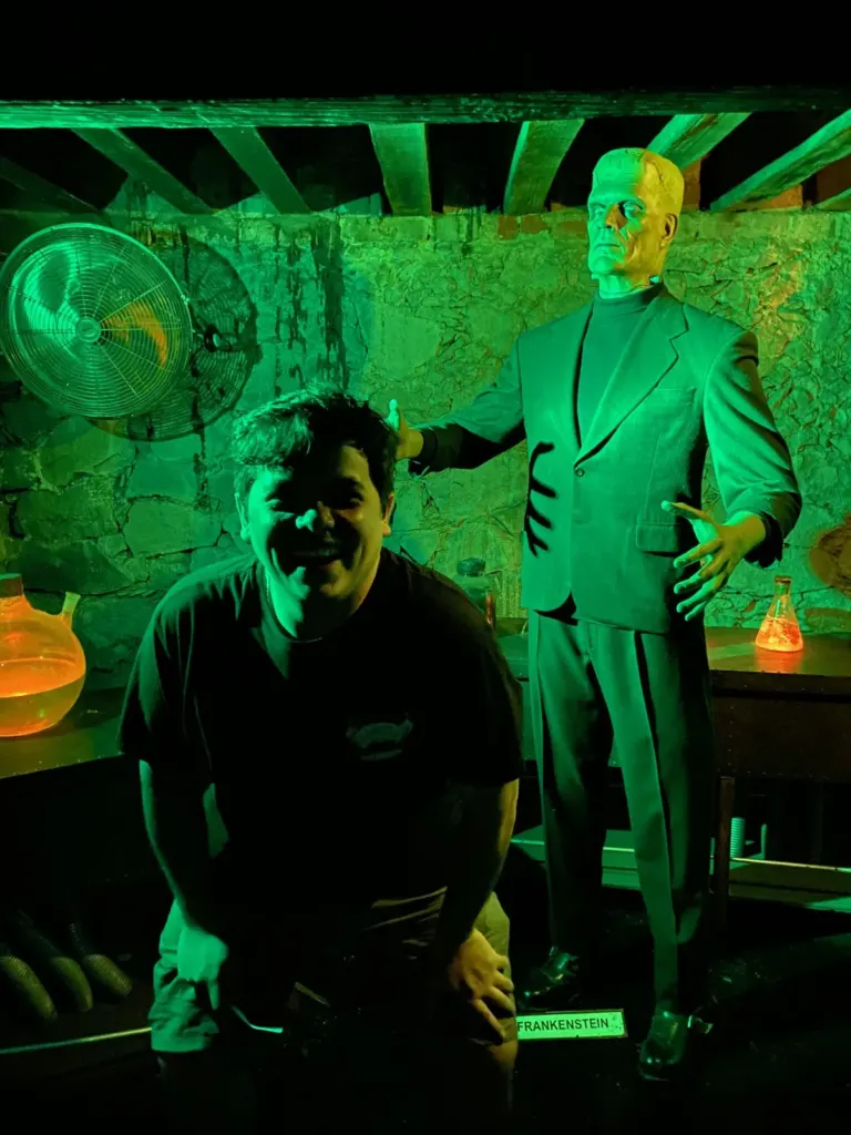 Figura de cera de Frankenstein con traje verde en un laboratorio iluminado con luz verde, con matraces naranjas y un ventilador viejo, y un visitante agachado al frente