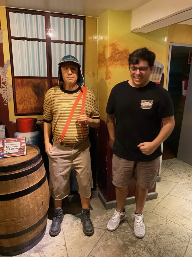 Figura de cera de El Chavo del Ocho con gorra, playera de rayas y tirante, junto a un barril de madera con su placa, y un visitante a su lado