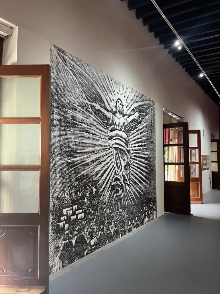 Grabado en blanco y negro ampliado a todo un muro, con una figura alada rodeada de rayos sobre una multitud, junto a puertas de madera del museo