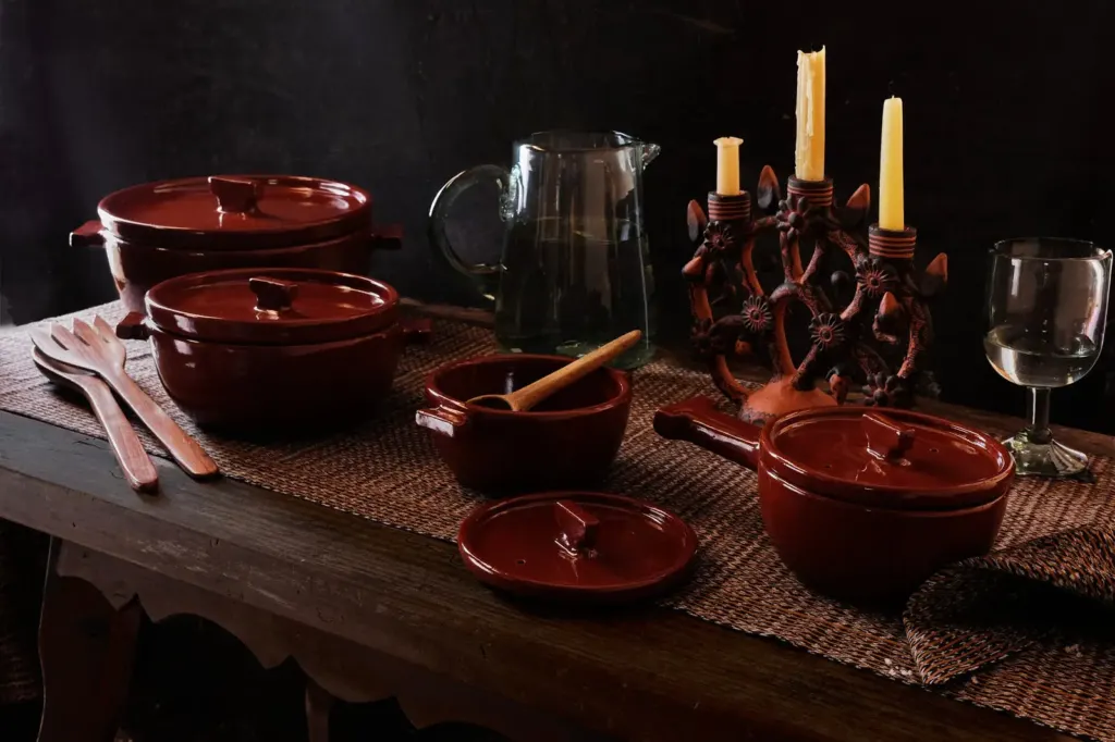 Bodegón de la colección Arrieta de Onora: cazuelas de barro vidriado en rojo oscuro, una jarra de vidrio, un candelabro de madera con velas y una copa sobre una mesa de madera, sobre fondo negro