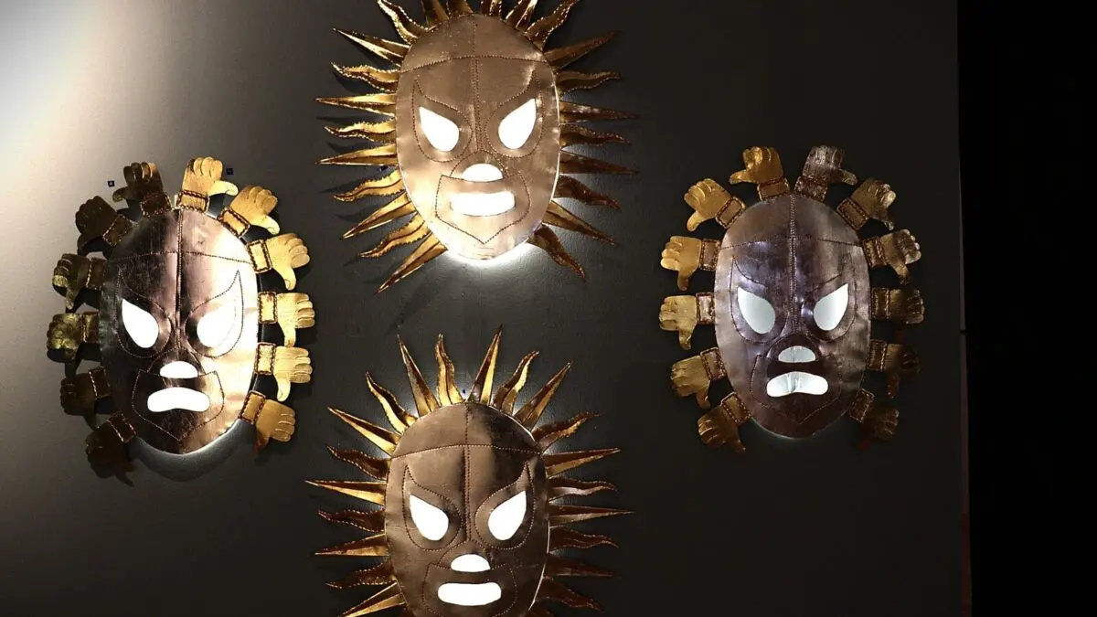 Cuatro máscaras de lucha libre hechas en metal dorado y cobre, montadas sobre un muro oscuro, obra de Betsabeé Romero