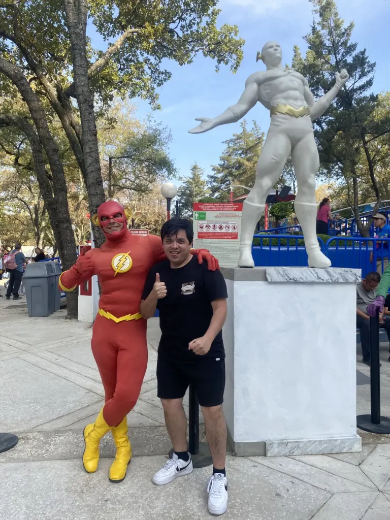 Hombre posando con un personaje de Flash junto a una estatua blanca de Flash sobre un pedestal en Six Flags México